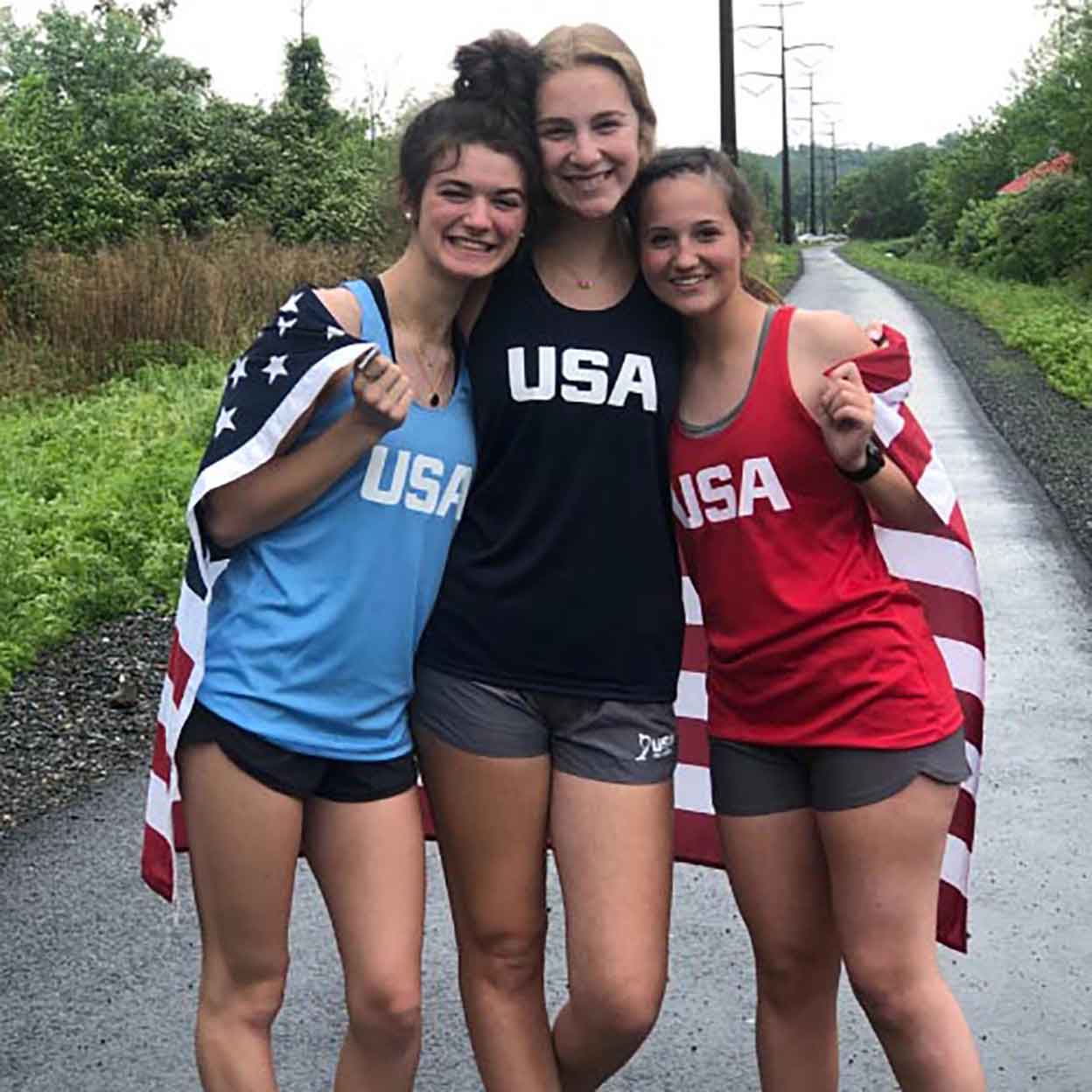Three smiling girls wearing the USA Field Hockey Performance Tanks with a flag around their backs.