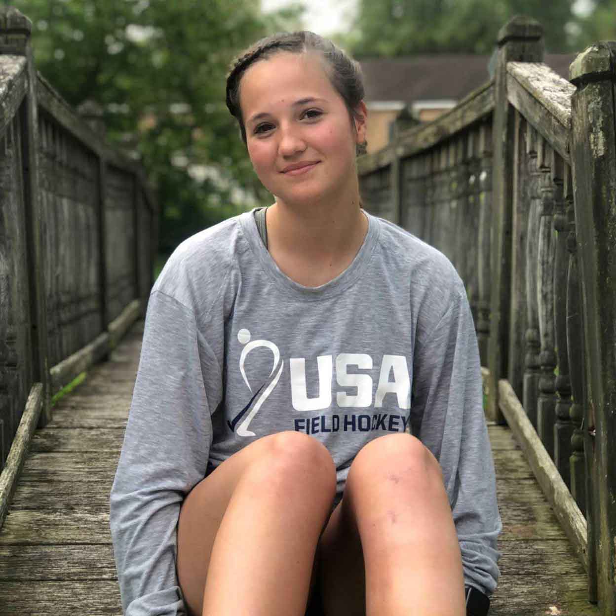Girl sitting on a bridge wearing the grey USA Field Hockey Long Sleeve Performance Tee