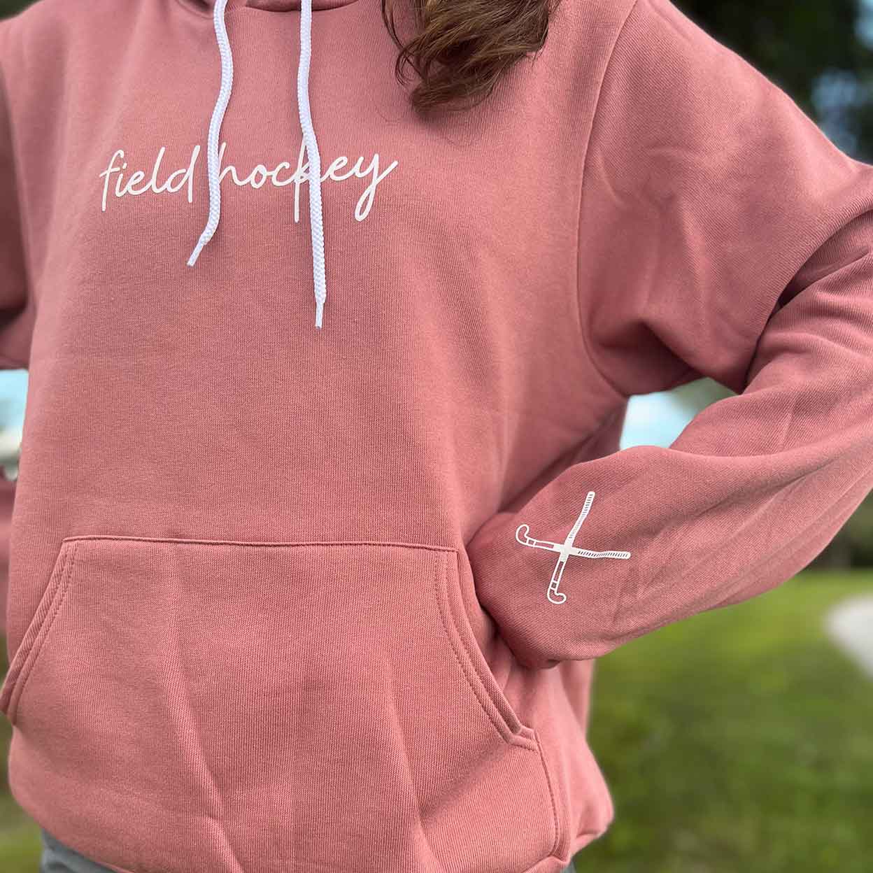 model wearing the pink Field Hockey Unisex Pullover Hooded Sweatshirt with hand on hip showing the crossed sticks on the wrist.