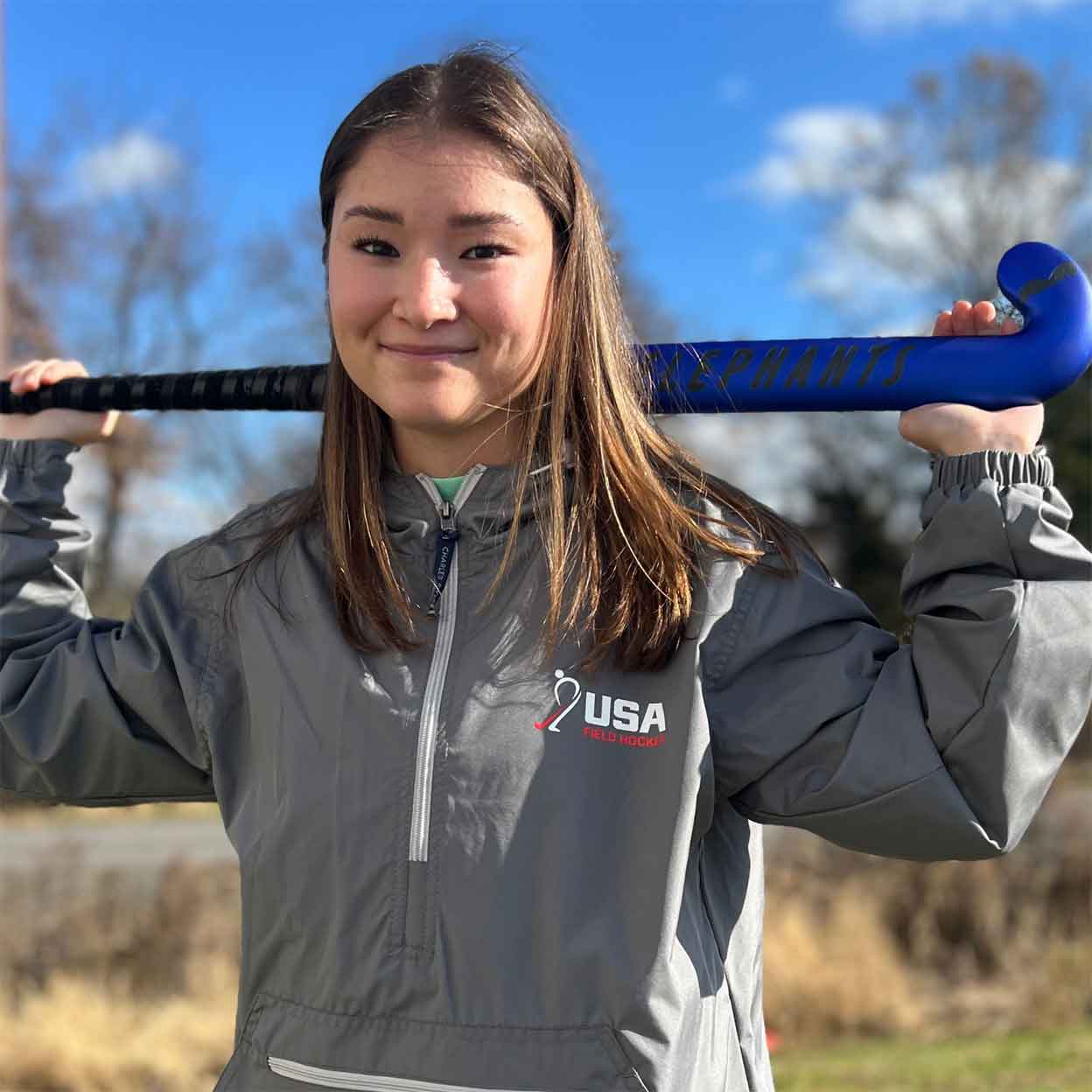 Smiling girl wearing the USA Field Hockey Unisex Anorak Jacket holding a field hockey stick behind her neck.