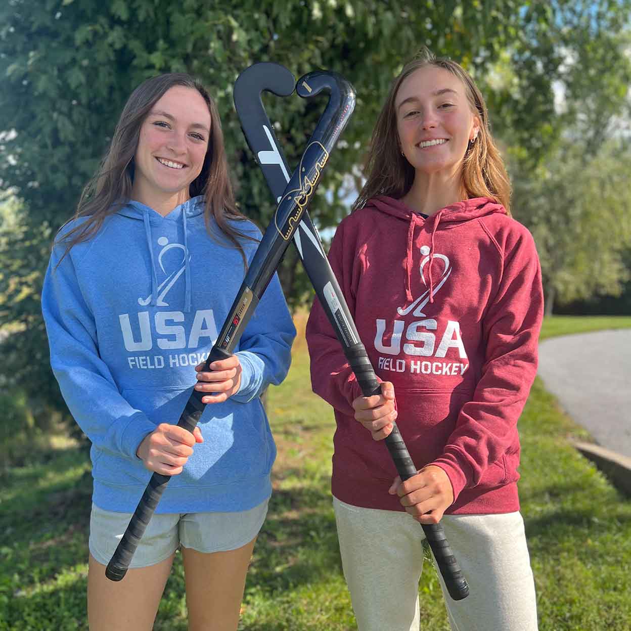 Two girls holding field hockey sticks and wear the USA Field Hockey Unisex Raglan Hooded Sweatshirts