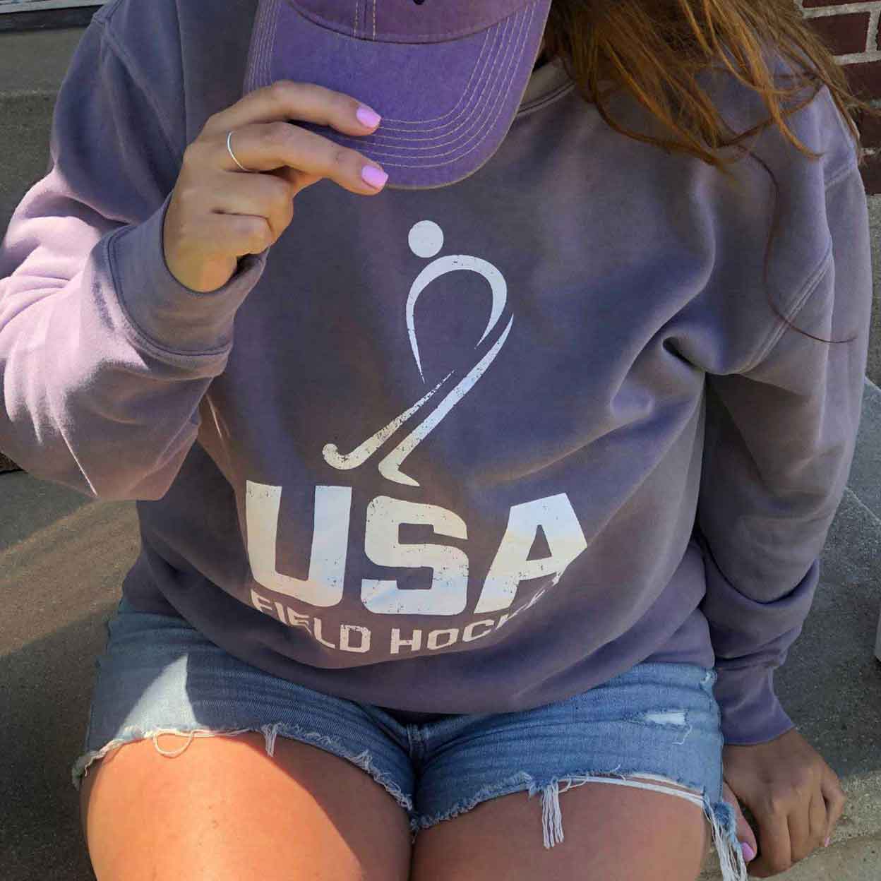 Girl wearing the USA Field Hockey Pigment-Dyed Crewneck Sweatshirt in purple with a purple hat. Her hand is holding onto the hat.